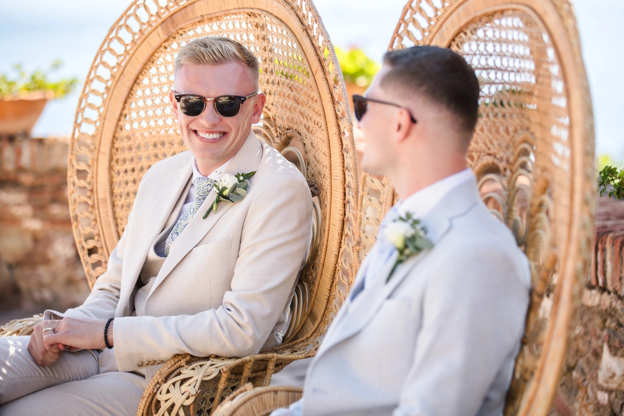Two grooms sit facing each other in woven peacock chairs, holding hands and smiling during their wedding ceremony. — Wedding at Hotel Only You, Malaga, spain
