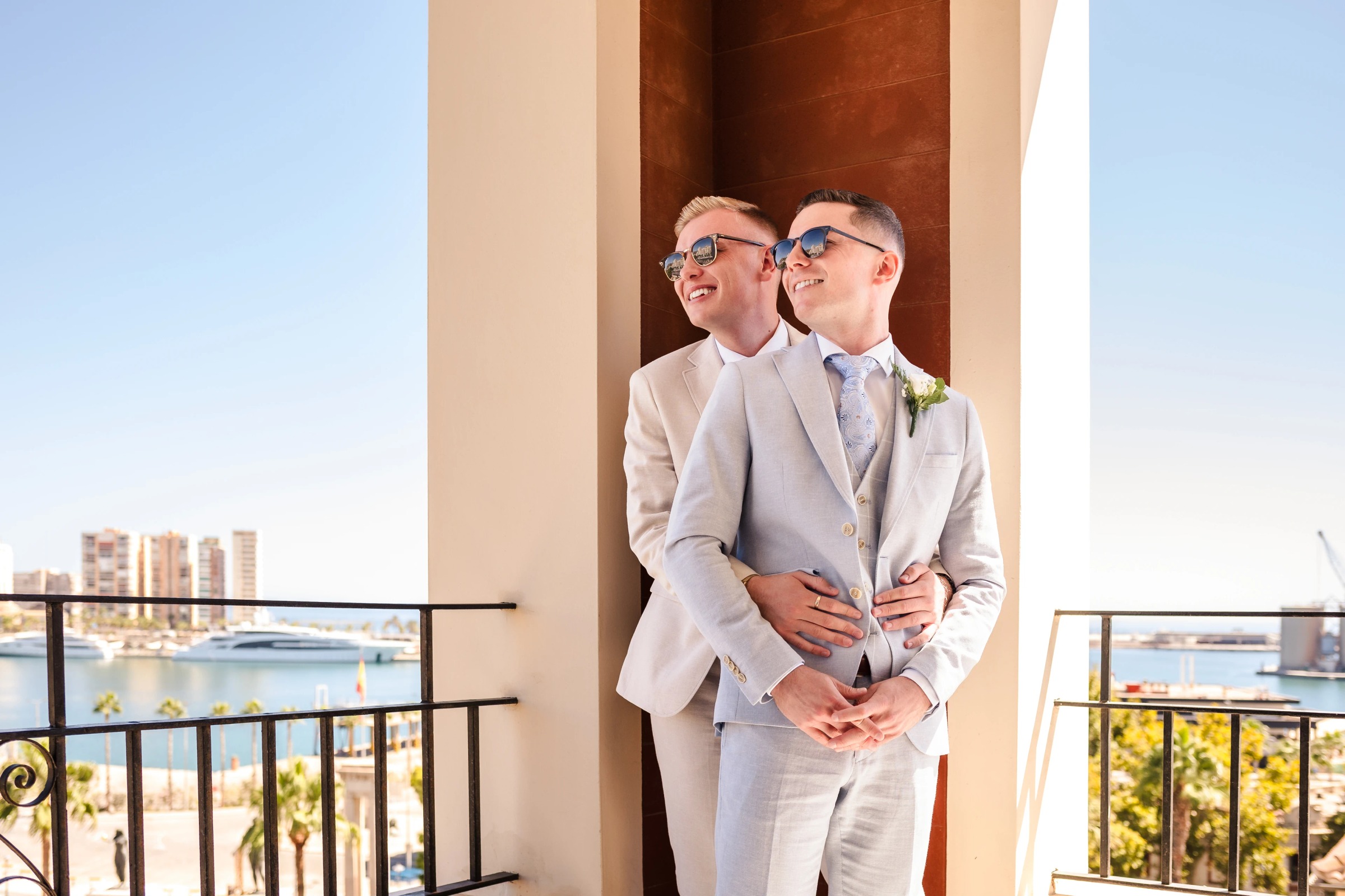 Two men in light suits embrace on a high-rise balcony overlooking a city, sharing an intimate moment. — Wedding at Hotel Only You, Malaga, spain