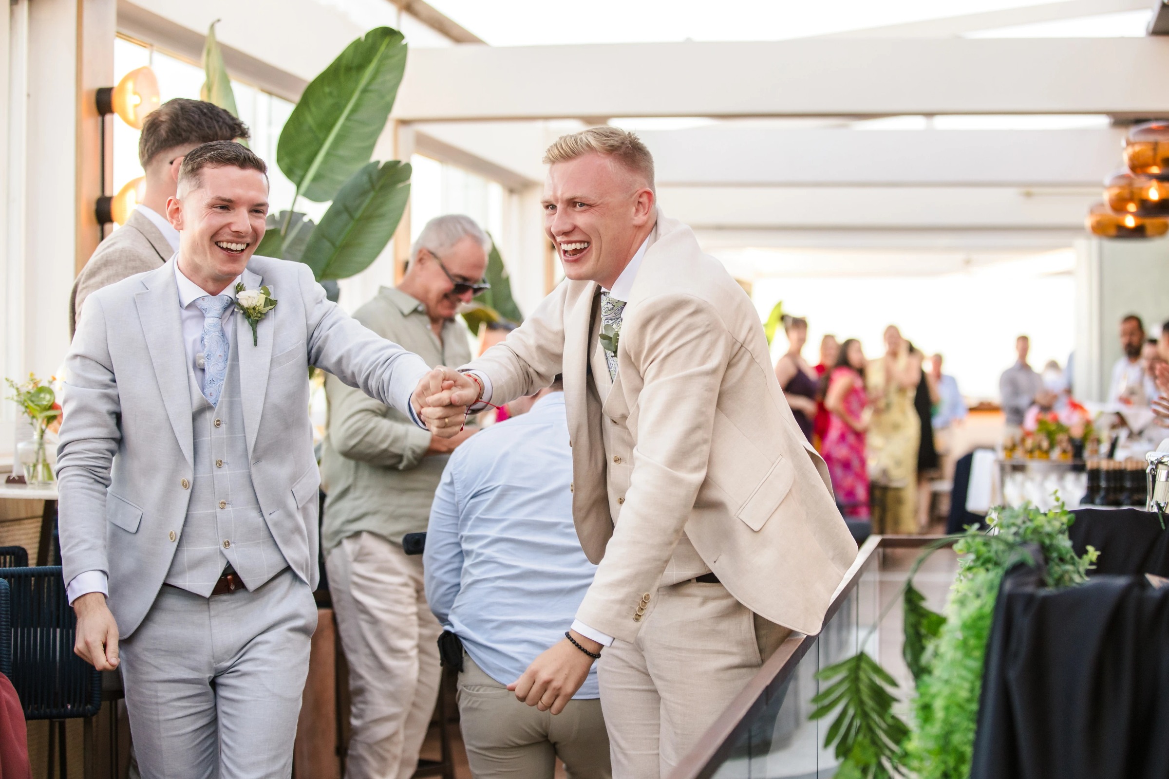 Two men in suits laugh and shake hands at a wedding reception while guests mingle in the background. — Wedding at Hotel Only You, Malaga, spain