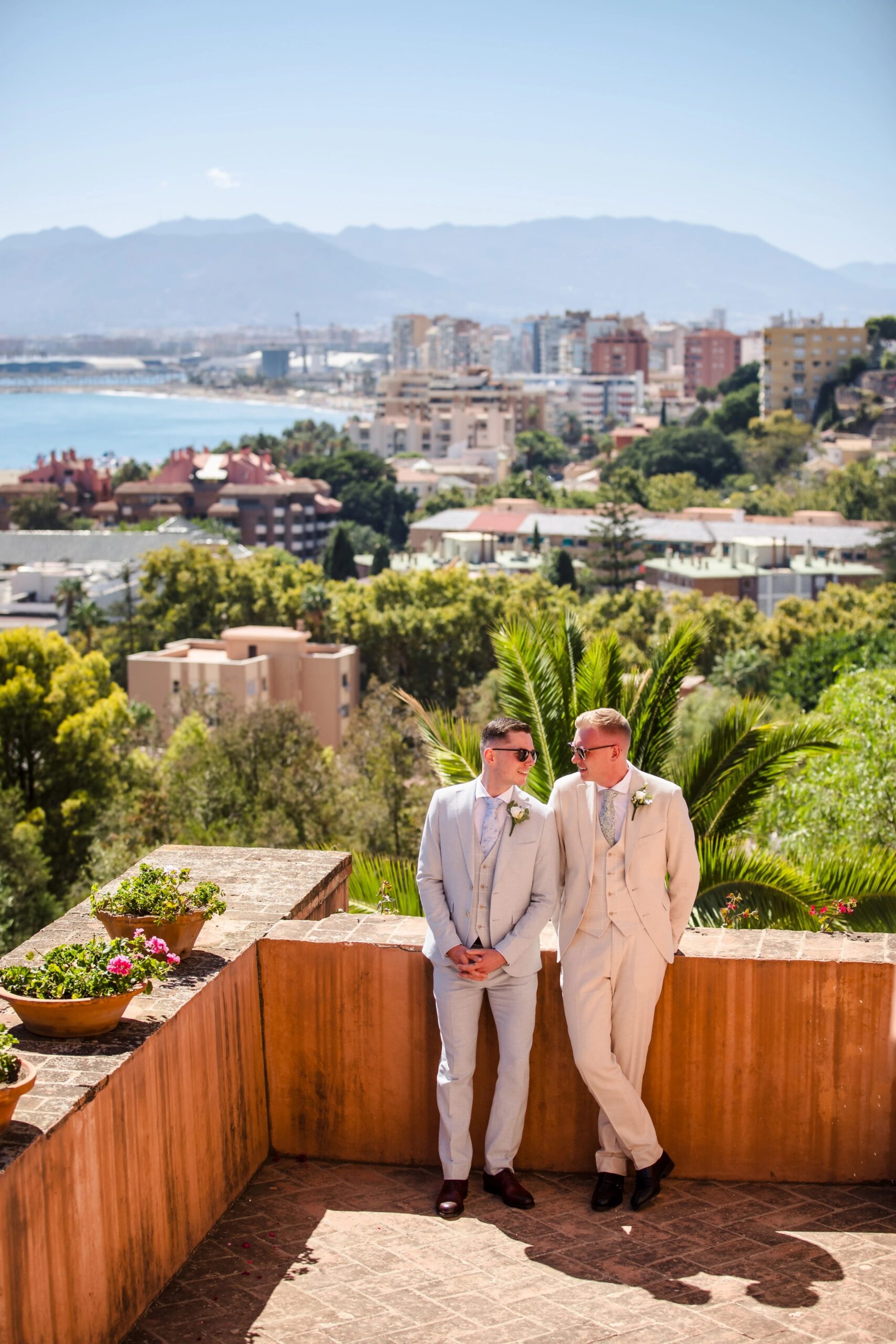 Two men in suits stand together on a terrace overlooking a coastal city, posing for a formal portrait. — Wedding at Hotel Only You, Malaga, spain